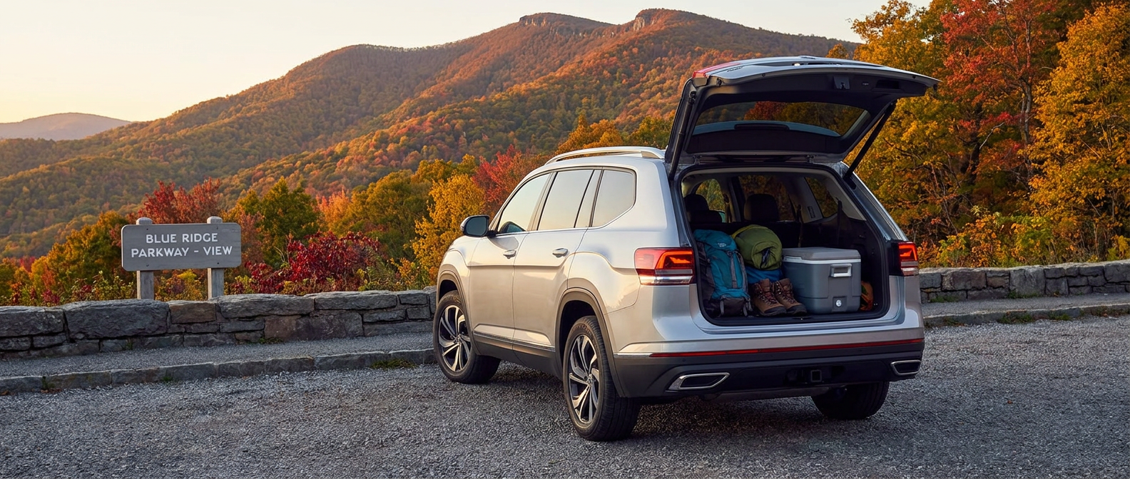 A 2026 Volkswagen Atlas in silver or deep blue parked at a scenic Blue Ridge Parkway overlook in North Carolina, rear liftgate open revealing packed weekend hiking gear, autumn foliage filling the mountain backdrop, afternoon light, shot from a low angle emphasizing the ridgeline.